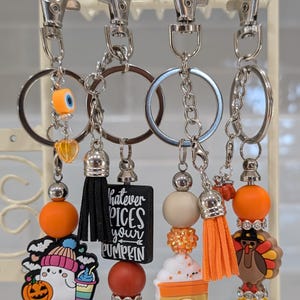 May include: Four keychains with silver hardware and decorative beads. Each keychain features a different charm, including a ghost with a pumpkin, a black tassel, a coffee cup, and a turkey. The keychains have orange, pink, and white beads.