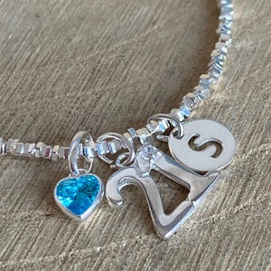 Op de afbeelding: Zilveren armband met meerdere bedels. Inclusief een blauwe hartvormige edelsteen, het nummer 21 en een ronde bedel met de letter "S". De armband ligt op een houten oppervlak.