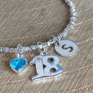 May include: A silver bracelet with a beaded chain and multiple charms. Charms include a blue heart-shaped gem, the number 18, and a letter "S". The bracelet is displayed on a wooden surface.