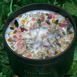 May include: A black candle in a round container, topped with white wax, colourful gemstones and dried herbs. The candle has a single wick and the text "Lotus Moon" is printed on the side.