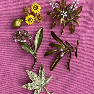 May include: A selection of floral and leaf brooches. The brooches include designs of yellow sunflowers, white flowers, and green leaves, some with pearl accents. The brooches are displayed on a pink background.