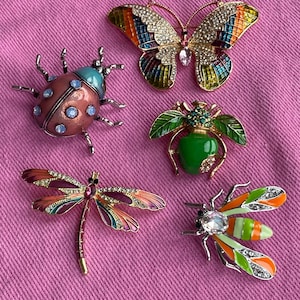 May include: Five insect brooches, including a ladybug, butterfly, dragonfly, bee, and beetle. Each brooch is made with colourful enamel and rhinestone detailing. The brooches are displayed on a pink fabric background.