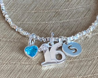 May include: A silver bracelet with multiple charms. Charms include a blue heart, the number 16, and a disc with the letter "S". The bracelet is made of small, square silver beads.