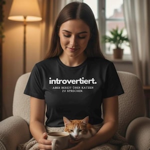 Könnte beinhalten: Schwarzes T-Shirt mit dem deutschen Text "introvertiert. Aber bereit über Katzen zu sprechen." in Weiß. Die Person trägt das Shirt und hält eine Tasse und eine Katze.