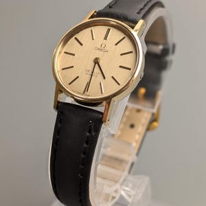 May include: A gold-toned Omega De Ville Quartz watch with a black leather band. The watch face is a light gold color with black hour markers and hands. The watch is displayed on a clear stand.