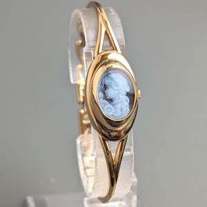 May include: A gold-tone bangle watch featuring an oval-shaped face with a light blue cameo inlay. The cameo depicts a profile of a person surrounded by floral details. The watch has a polished metal finish and a split-shank band design.