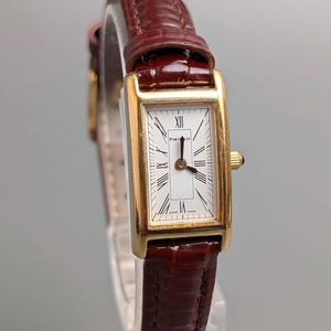 Può includere: Un orologio da polso rettangolare dorato con quadrante bianco e numeri romani. L'orologio ha un cinturino in pelle marrone. Il quadrante dell'orologio indica "Porto 10" e "Swiss Made".
