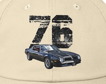 Może przedstawiać: Zniszczona, beżowa czapka z daszkiem z czarną grafiką klasycznego samochodu i numerem "76" w zniszczonej czcionce. Czapka ma wstępnie zakrzywiony daszek i zużytą, vintage estetykę.