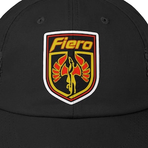 May include: Black distressed baseball cap featuring a shield-shaped emblem with the word "Fiero" in yellow. The emblem includes a stylized bird in red and yellow, set against a white and black background. The cap has a curved brim.