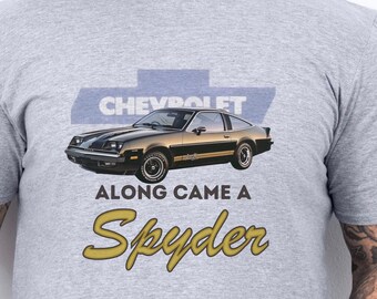 Może przedstawiać: Jasnoszara koszulka z grafiką klasycznego samochodu Chevrolet Spyder w kolorze czarnym i złotym. Tekst "CHEVROLET" znajduje się nad samochodem, a "ALONG CAME A Spyder" pod nim. Koszulka wykonana jest z miękkiego materiału.