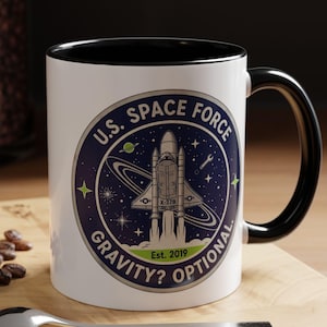 Puede incluir: Taza de cerámica blanca con interior y asa negros. La taza presenta un emblema de la Fuerza Espacial de EE. UU. con un gráfico de transbordador espacial, el texto "Gravity? Optional" y el año "Est. 2019."