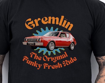Może przedstawiać: Czarny T-shirt z grafiką czerwonego samochodu AMC Gremlin z tekstem "Gremlin The Original Funky Fresh Ride" w kolorze pomarańczowym i żółtym.