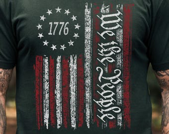 Może przedstawiać: Ciemnozielona koszulka z postarzanym wzorem amerykańskiej flagi. Flaga zawiera tekst "1776" w okręgu gwiazd i słowa "We the People". Koszulka ma krótkie rękawy.
