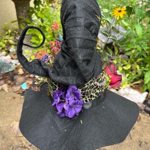 May include: A black felt witch hat featuring a tall, pointed crown with a curled tip. The hat is decorated with a wide band of gold-toned netting, a cluster of vibrant purple silk flowers, and various dried floral accents tucked into the brim.