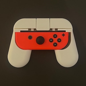 Könnte beinhalten: Weißer Kunststoff-Controller-Griffaufsatz für einen Nintendo Switch Joy-Con-Controller. Der Griff hat zwei weiße Griffe und einen roten Joy-Con-Controller in der Mitte.