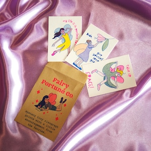 Puede incluir: Tres tarjetas rectangulares pequeñas con ilustraciones coloridas de hadas y texto, acompañadas de un sobre de papel marrón etiquetado Fairy Fortune Co. con un gráfico de hada y el texto Mystery tiny risograph prints with a little encouragement.