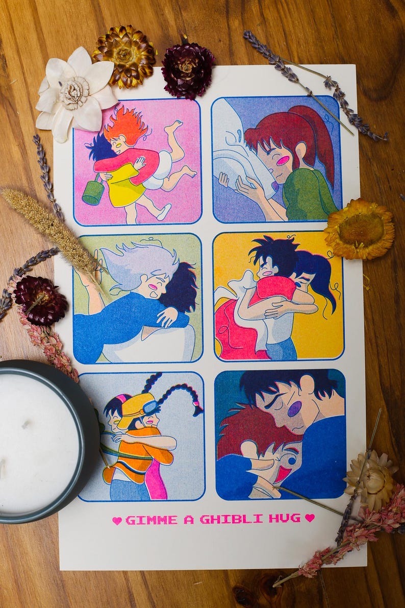 May include: A white poster with six colorful panels depicting animated characters hugging. The phrase "GIMME A GHIBLI HUG" is printed at the bottom in pink. The poster is surrounded by dried flowers and a candle.