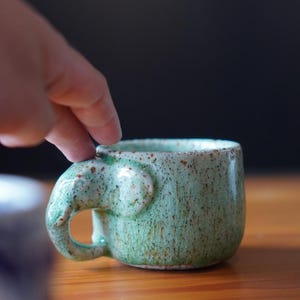 May include: A handmade ceramic mug with an elephant ear handle. The mug has a speckled, light green glaze with brown accents. The mug is on a wooden surface.