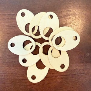 Könnte beinhalten: Eine Ansammlung ovaler Holzanhänger mit einem kreisförmigen Loch und einem kleineren Bohrloch. Die Anhänger sind hellbeige und kreisförmig auf einer dunkelbraunen Holzoberfläche angeordnet.