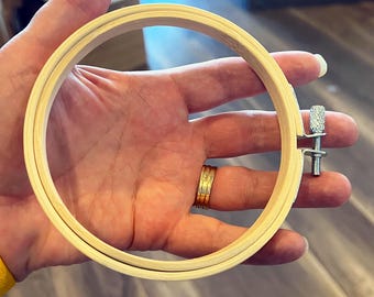 May include: A wooden embroidery hoop with a metal tightening screw. The hoop is held in a hand.