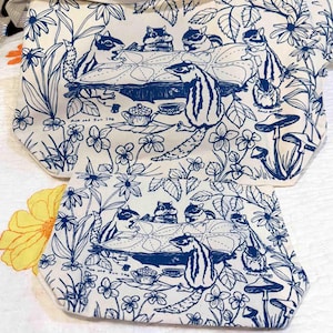 Puede incluir: Juego de bolso tote y posavasos blanquecino con una ilustración azul de ardillas listadas en una mesa, rodeadas de flores y follaje. El texto "Mum and Sun Ink" es visible. El bolso tote tiene una correa color crema.