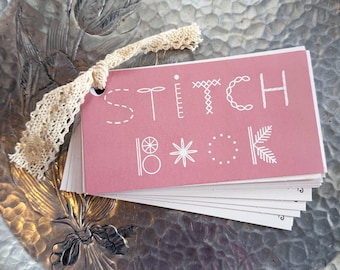 Może przedstawiać: Różowa książeczka z napisem "STITCH BOOK" napisanym na biało z różnymi wzorami ściegów. Książeczka jest związana beżową wstążką z koronki.