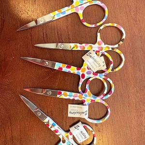 May include: Five pairs of colorful embroidery scissors with white handles and polka dot patterns. The scissors have a label that reads "Embroidery Scissors Item No. 6340-10".