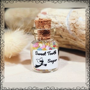 May include: A small glass jar with a cork stopper filled with colorful sugar crystals. The label reads "Sweet Tooth Sugar" and features a small fairy illustration.