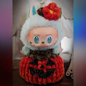 May include: A whimsical doll with a light blue furry hood and bunny ears, adorned with a red flower. The doll is seated on a plush, orange pumpkin with a carved jack-o'-lantern face. The doll's eyes are large and teal.