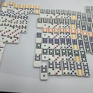 May include: A collection of white dominoes with colored dots arranged on a white surface. The dots are in various colors, including red, blue, green, orange, and yellow. The dominoes are arranged in a pattern.