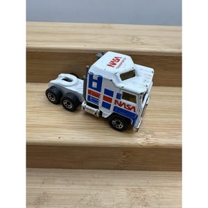 Pode incluir: Um brinquedo de semi-reboque branco e azul com o logotipo da NASA e a palavra "TRANSPORTES" em vermelho. O caminhão tem rodas pretas e um engate de reboque. O brinquedo está em uma superfície de madeira.