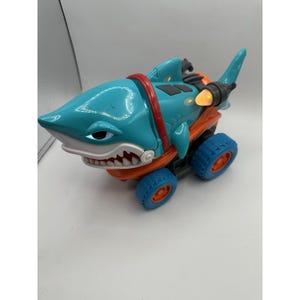 May include: A toy vehicle shaped like a shark with a teal body, orange undercarriage, and blue wheels. The shark features a red ring around its midsection, a white mouth with sharp teeth, and a glowing orange light on the rear fin assembly.