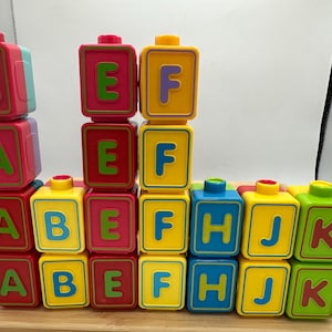 May include: Brightly coloured alphabet blocks in red, yellow, blue, and green. Each block displays a letter, with letters A to K visible. The blocks are stacked on a wooden surface, creating a playful learning environment.