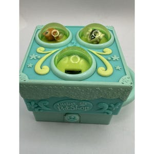 Puede incluir: Un juguete de Littlest Pet Shop de color turquesa con tres cúpulas transparentes que contienen pequeñas figuras de animales. El juguete tiene un cajón extraíble y detalles decorativos florales y de estrellas. El texto "Littlest Pet Shop" es visible en la parte delantera.