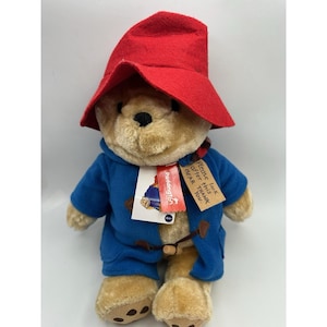 May include: A plush Paddington Bear wearing a red felt hat and a blue coat. The bear has a tag that says "Please look after this bear. Thank you."