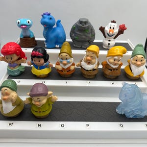 Può includere: Una collezione di figurine colorate di personaggi Disney, tra cui una sirena, Biancaneve, nani e personaggi di Frozen. Le figurine sono disposte su un espositore a più livelli, mostrando una varietà di personaggi animati.