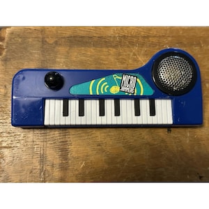 Op de afbeelding: Een blauw speelgoedkeyboard met witte en zwarte toetsen, een zwarte luidspreker en de woorden "Micro Jammers" op de voorkant. Het keyboard heeft een zwarte knop en een turquoise en geel ontwerp.
