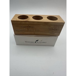 May include: A rectangular light brown bamboo wooden stand with three circular cutouts on top. The front is engraved with the text Young Living Natural Boost Bar and a Seed to Seal logo. It rests on a white cardboard box labeled Young Living Essential Oils.