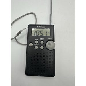 May include: A black RadioShack portable digital radio featuring a rectangular body, a digital display showing 105.1 MHz, a silver telescopic antenna, a grey wrist strap, and circular control buttons for power, programme, tuning, and AM/FM selection.