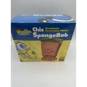 Puede incluir: Una caja azul con un Chia Pet de Bob Esponja. La caja muestra una maceta de color terracota con la forma de la cara de Bob Esponja, llena de brotes verdes. La caja también tiene los logotipos de Nickelodeon y Bob Esponja.