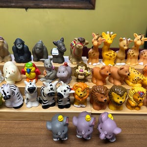 May include: A collection of colourful plastic animal figurines, including giraffes, zebras, elephants, lions, and monkeys. The figurines are arranged on a wooden shelf with three tiers.