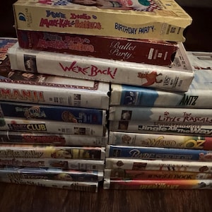 May include: A collection of VHS video tapes, including titles such as "Jumanji" and "Antz." The tapes are stacked, with the titles and graphics visible on the spines. The tapes are in various colors and designs.