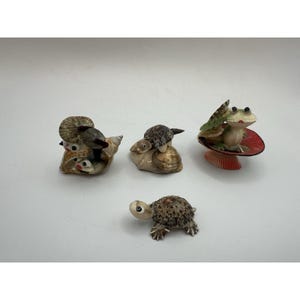 May include: A collection of small, decorative seashell figurines. The figurines include a turtle, a frog on a red lily pad, and several shell creatures. The shells are beige, brown, and white, with intricate details. The frog has green and white accents.