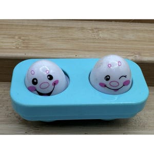 Può includere: Un giocattolo di plastica azzurra con due figure bianche a forma di uovo, ognuna con un volto sorridente, guance rosa e occhi neri. Una figura fa l'occhiolino. Giocattolo per bambini.
