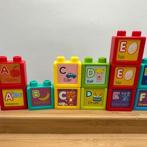 May include: Colorful alphabet blocks with letters A through F, each featuring an image and word. The blocks are red, yellow, green, and blue, with illustrations of an apple, airplane, car, doll, egg, and fawn.