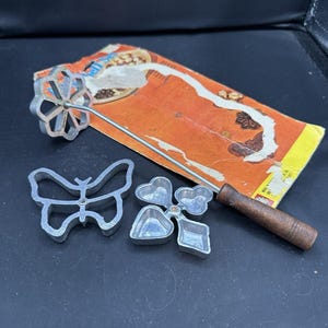 May include: A vintage rosette iron set with a wooden handle, a flower-shaped iron, and three other decorative molds. The set includes a butterfly, heart, and square shapes. The packaging is orange and yellow.
