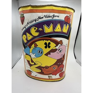 May include: Vintage Pac-Man metal wastebasket. The cylindrical bin features the Pac-Man logo and characters in a colorful design. The words "Midway's Amazing New Video Game" are printed on the side, along with images of cherries and a strawberry.