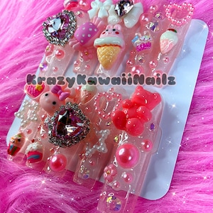 May include: Set of six pink and white resin hair clips with various kawaii charms, including a heart, a bunny, an ice cream cone, a cupcake, a bear, and a strawberry. The clips are decorated with pearls, glitter, and rhinestones. The text "Krazy Kawaii Nails" is visible on the bottom of the image.