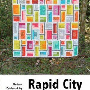 May include: A colourful quilt pattern called "Rapid City" by Elizabeth Hartman. The quilt features a patchwork design with geometric shapes in various colours, including red, orange, yellow, green, blue, and pink.