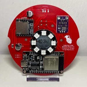 May include: A red circular circuit board featuring electronic components such as a speaker, memory card slot, and a purple sensor. The board is labelled "Juggernog Fridge Bluetooth Board v2.1" and "Designed by Infragno".